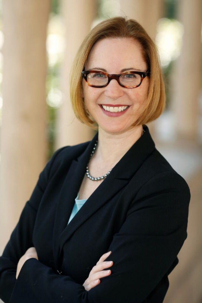 A woman in a black blazer with arms crossed, smiling. 