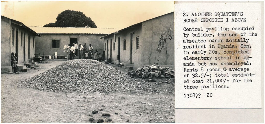 Austere one-story housing behind a dirt courtyard. People standing against the far wall. 